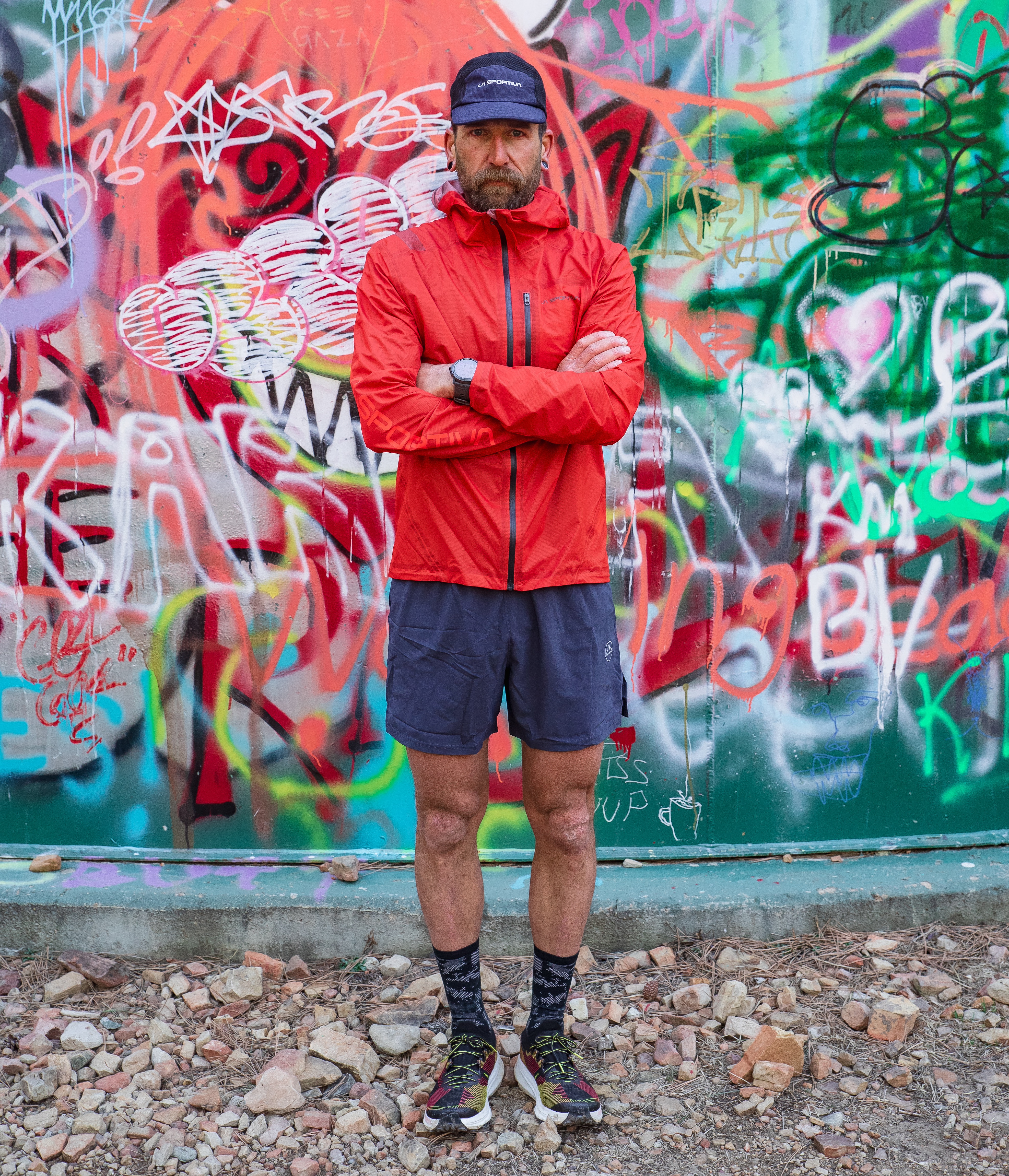 Man in red jacket stands with arms crossed in front of colorful graffiti wall.