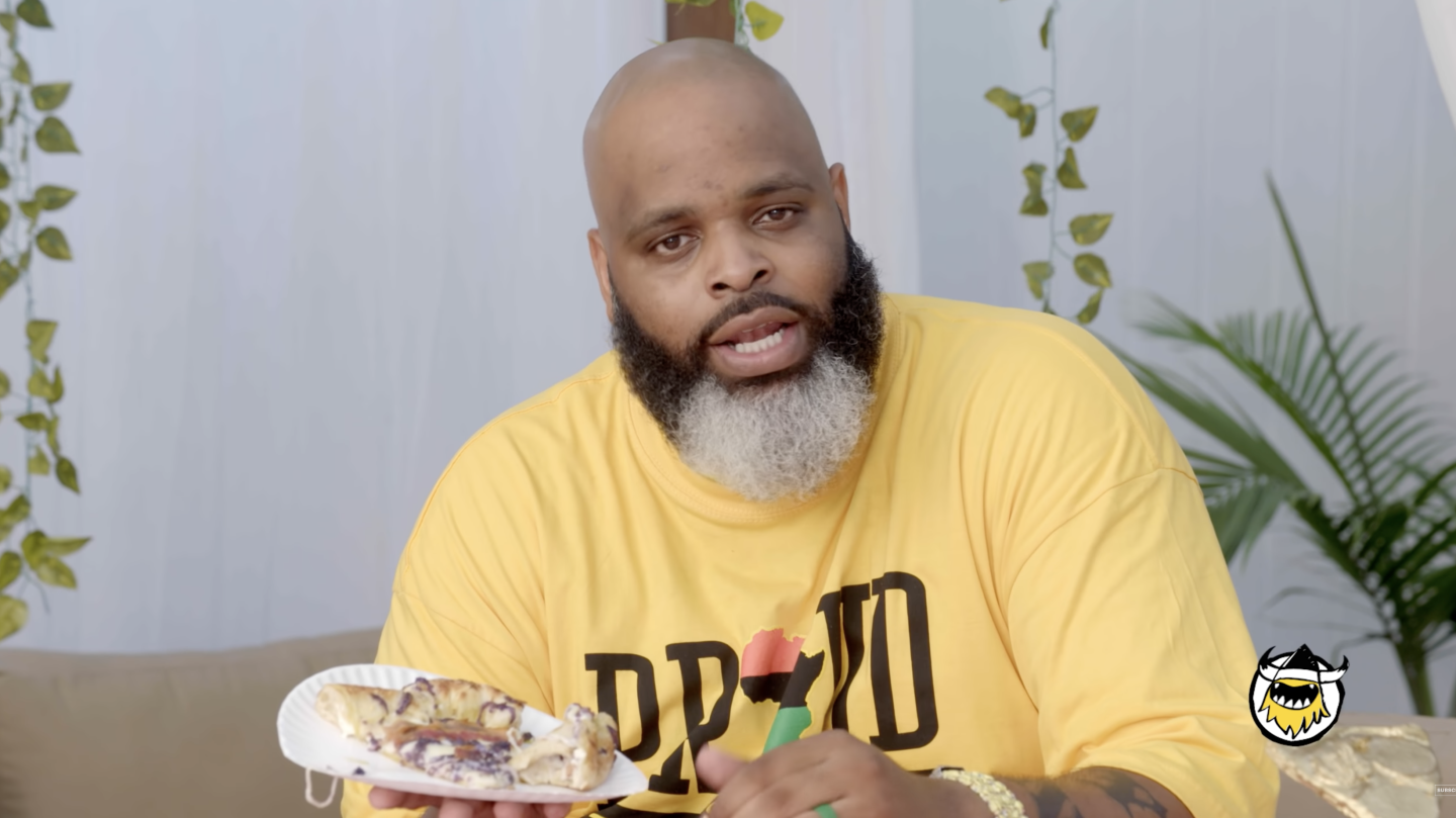 A man speaking to the camera holding a plate of pizza