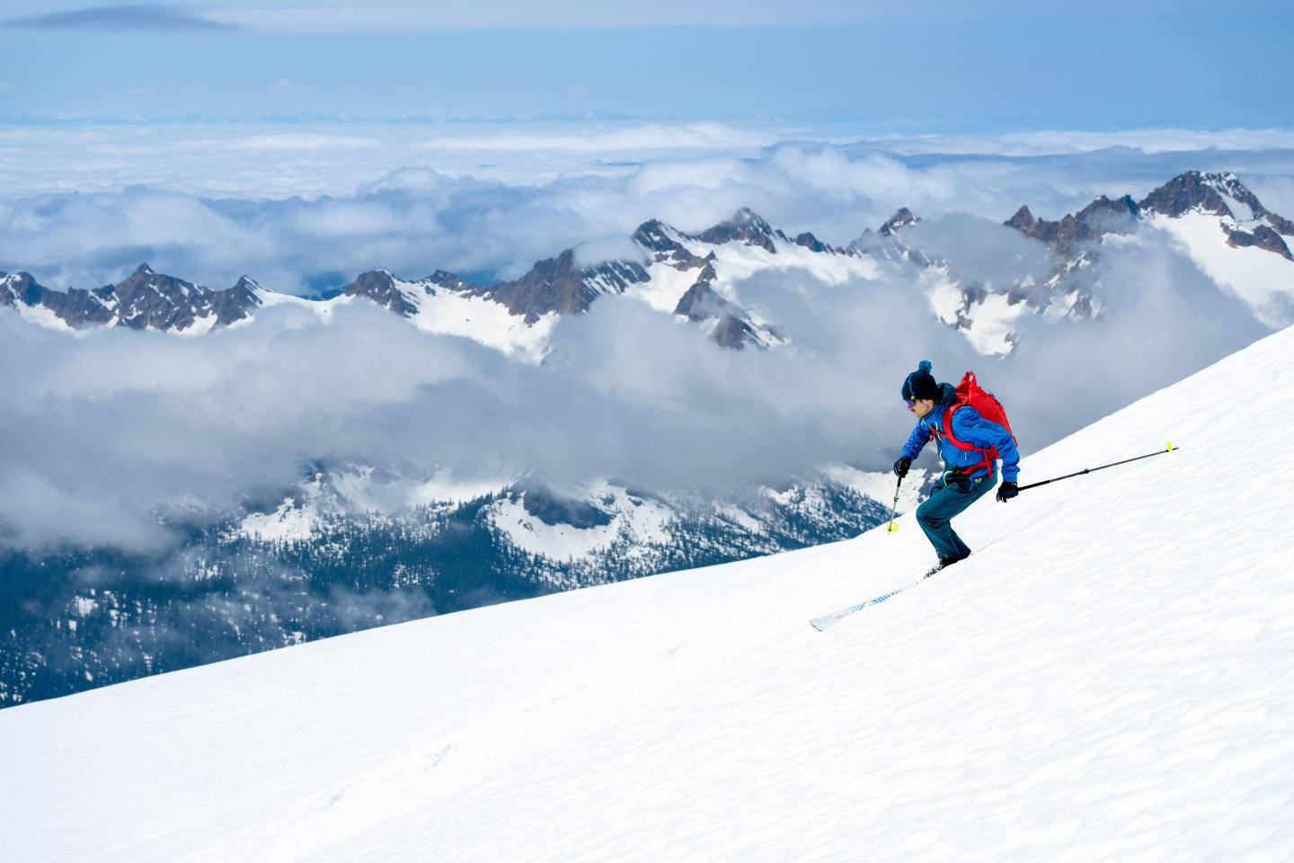 Man with red backpack is skiing down a steep snowy slope, fog covered mountains in the background