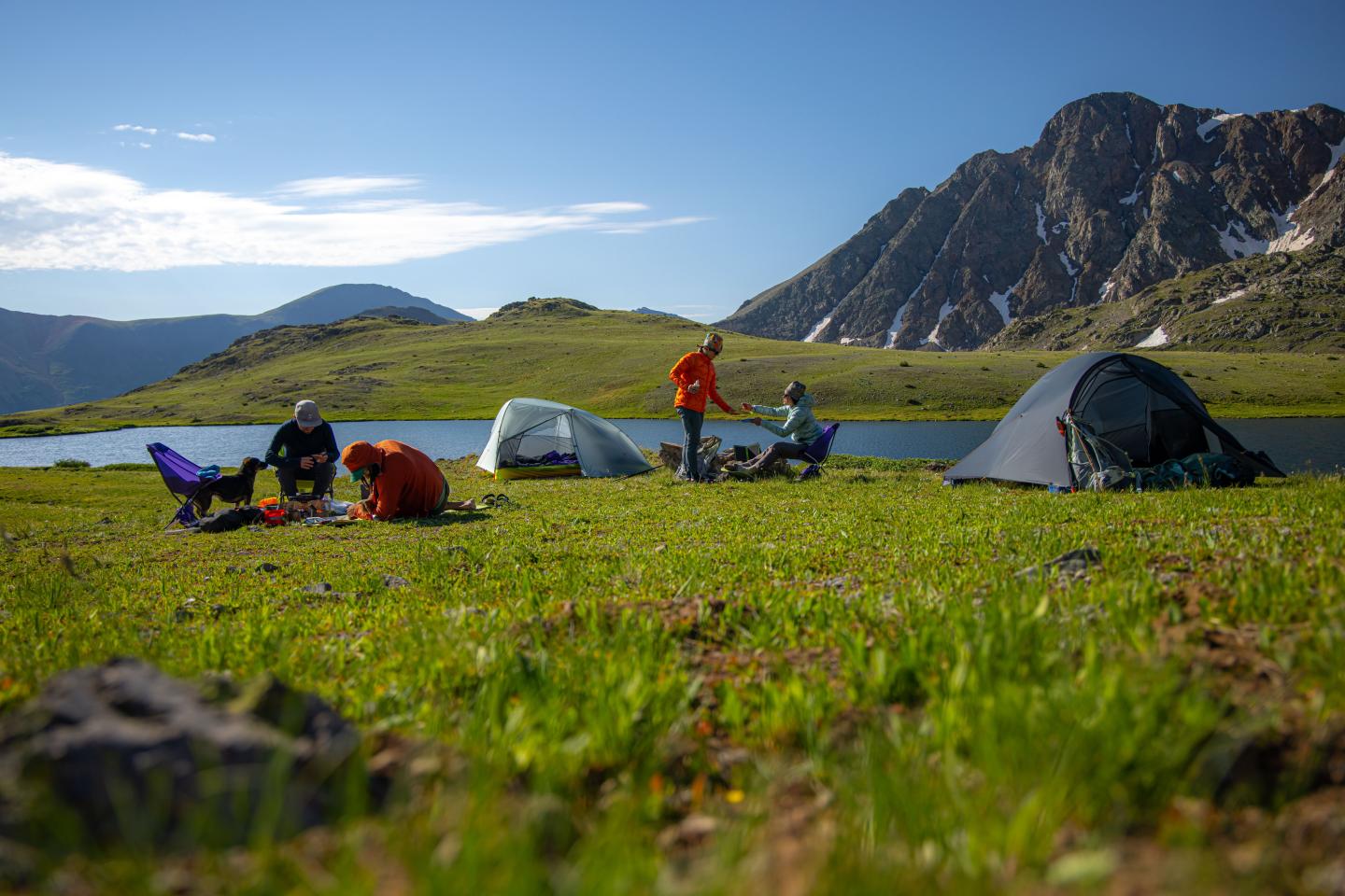 Campers setting up tents on a grassy lakeside, mountains in the background.
