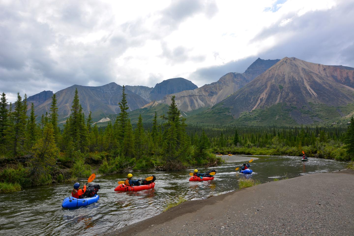 Kayakers in colorful boats navigate a river, surrounded by mountains and trees.