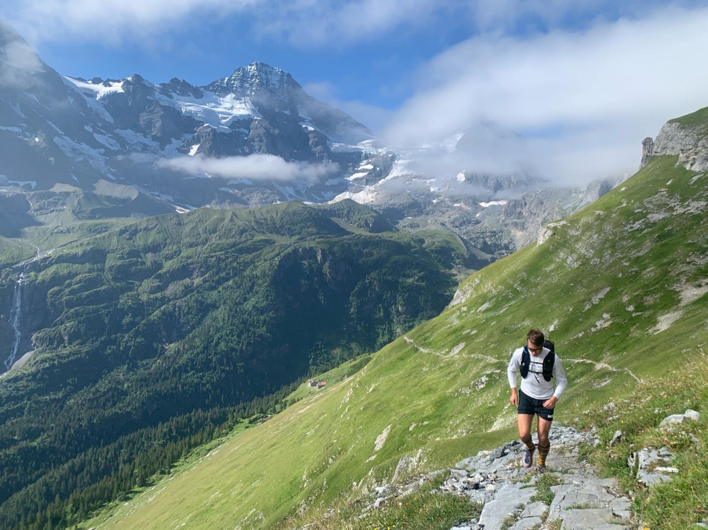Hiker on a mountain trail with green hills and snow-capped peaks in the background.