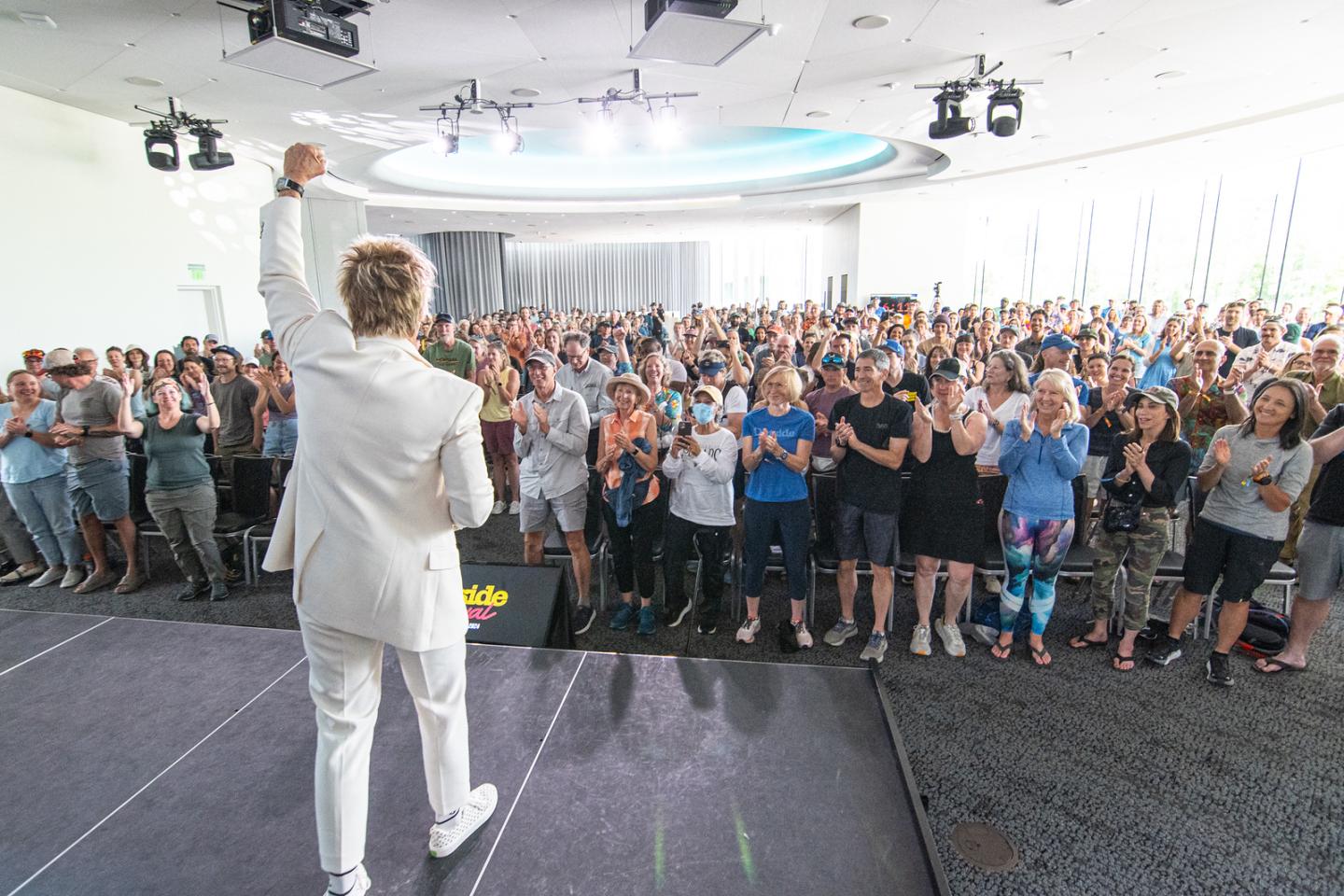 Person in white suit addresses large applauding audience in bright conference room.