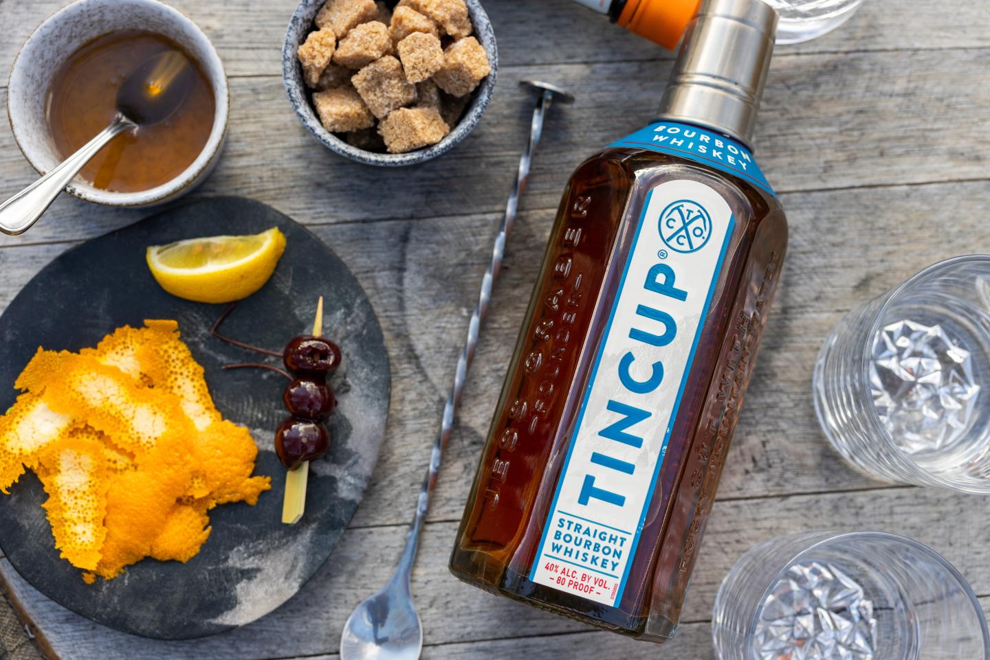 Tincup whiskey bottle next to a plate of orange peels, cherries, and sugar cubes on a table.