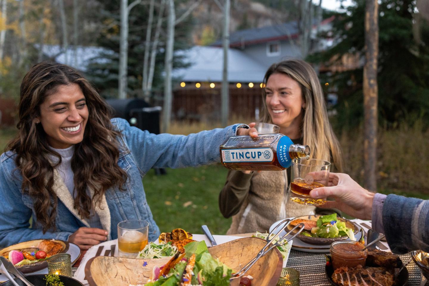 Friends sharing food and drinks at an outdoor table, smiling and laughing.