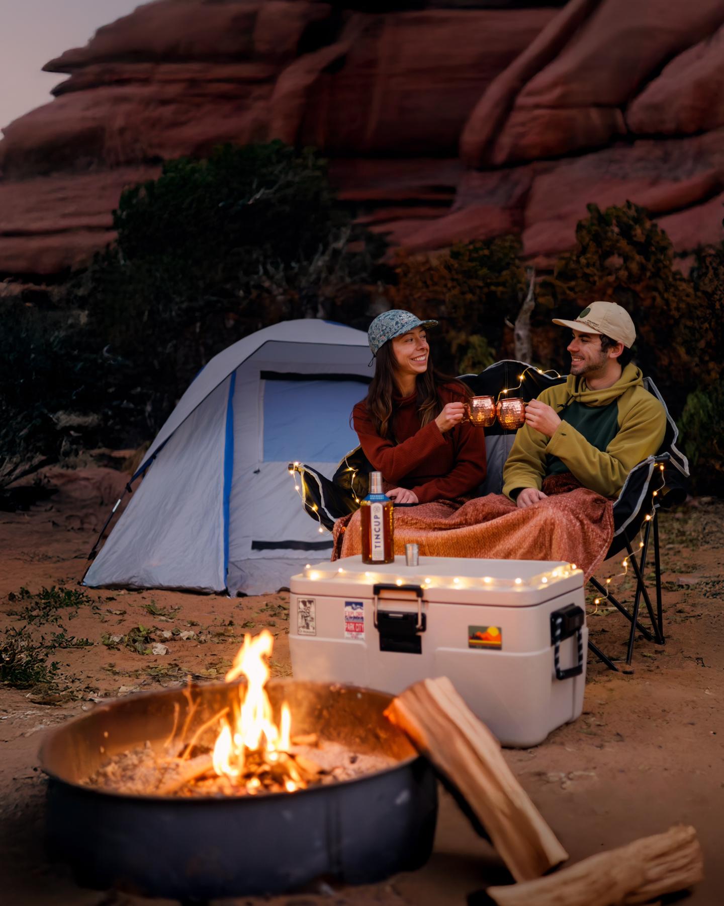 Campers toast by a fire in front of a tent and red rock formations.