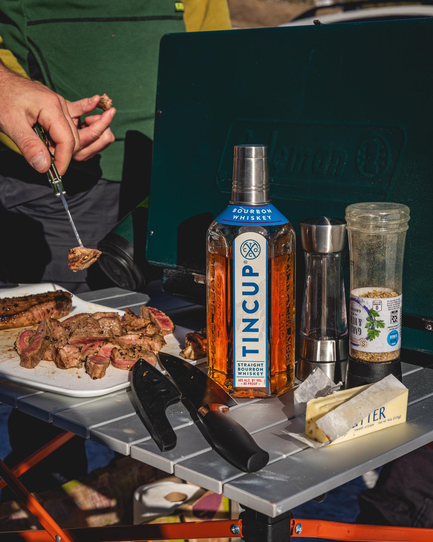Camping table with whiskey bottle, grilled steak, and cooking utensils.