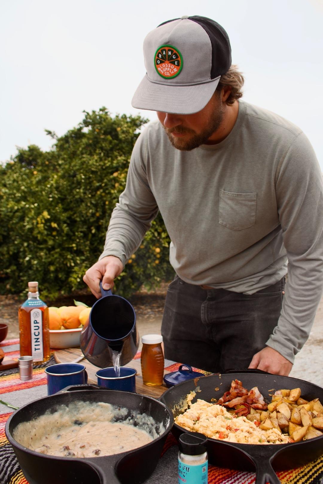 Man in cap pours drink at outdoor breakfast table with skillet meals.