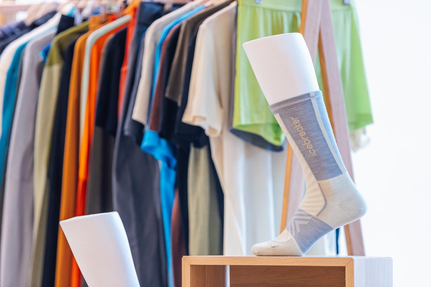 Sock on a mannequin leg with colorful clothes hanging in the background.