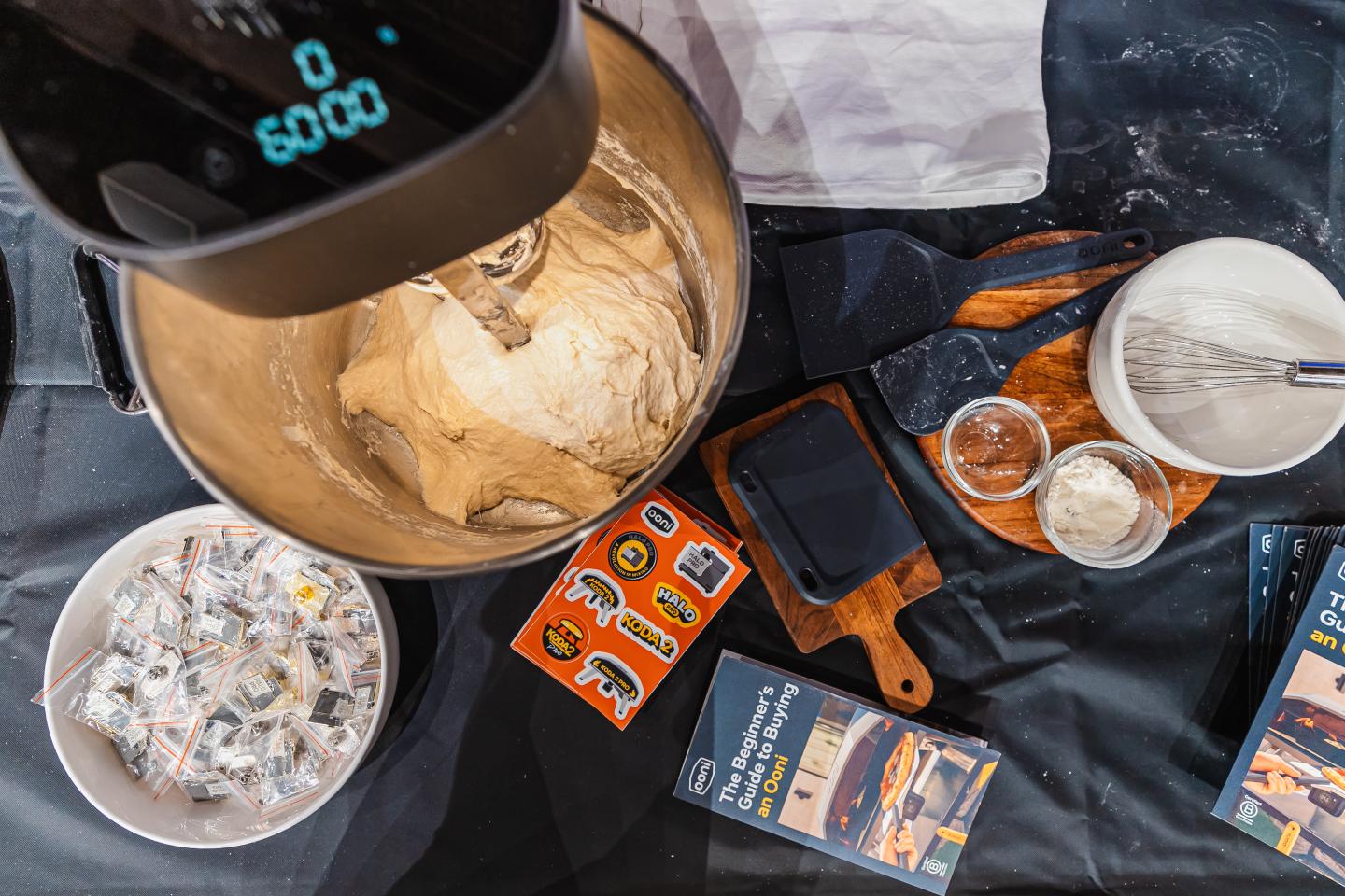 Mixer with dough, bowls, whisk, and recipe cards on a black tablecloth.