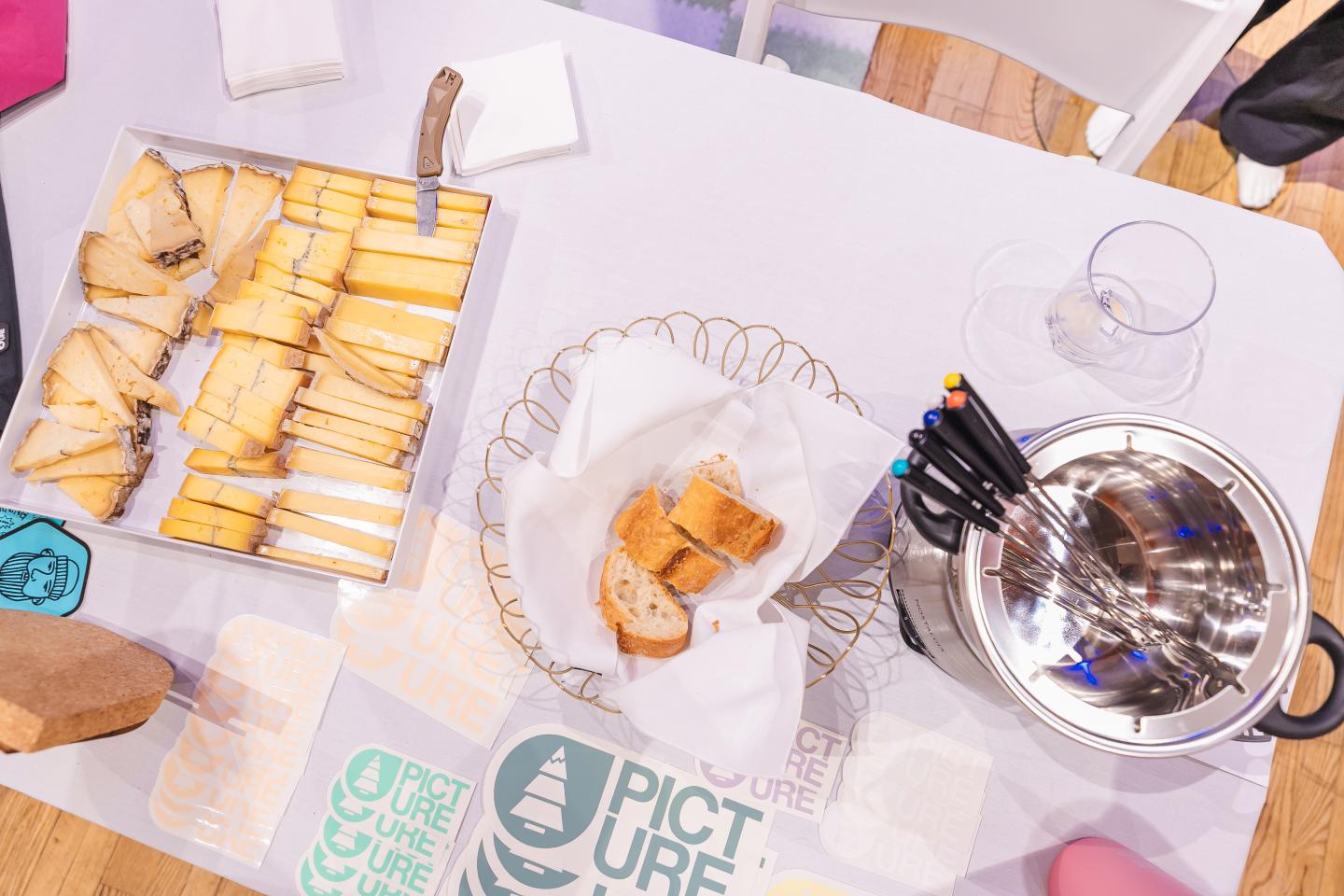 Bread tray, sliced cheese, and metal pot with pens on a white tablecloth.