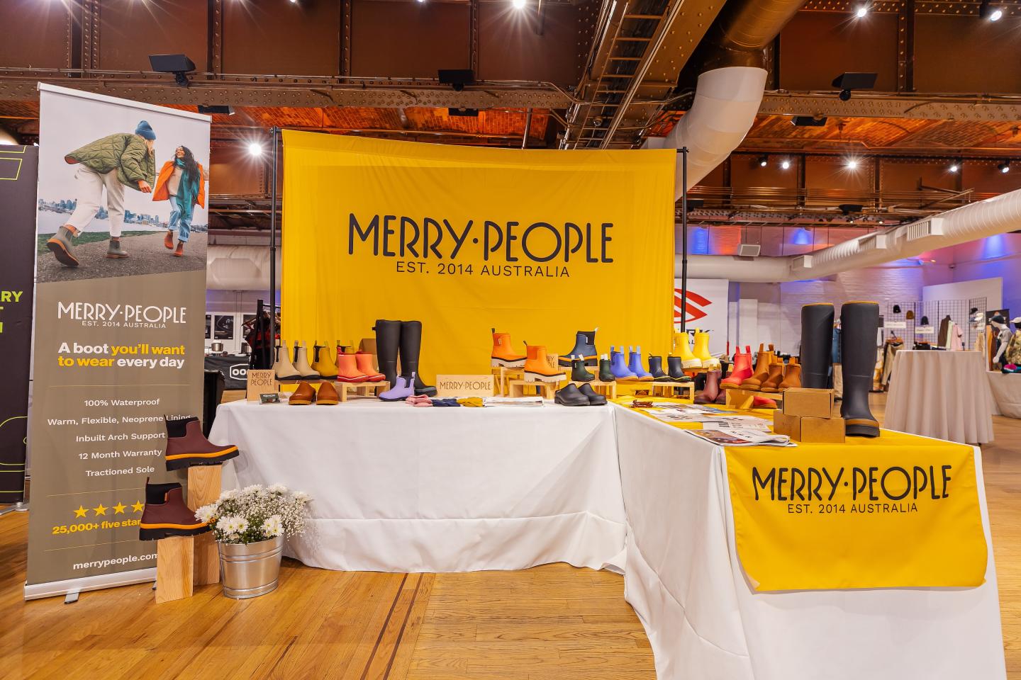 Trade show booth with bright yellow Merry People branding, displaying boots and promotional materials.