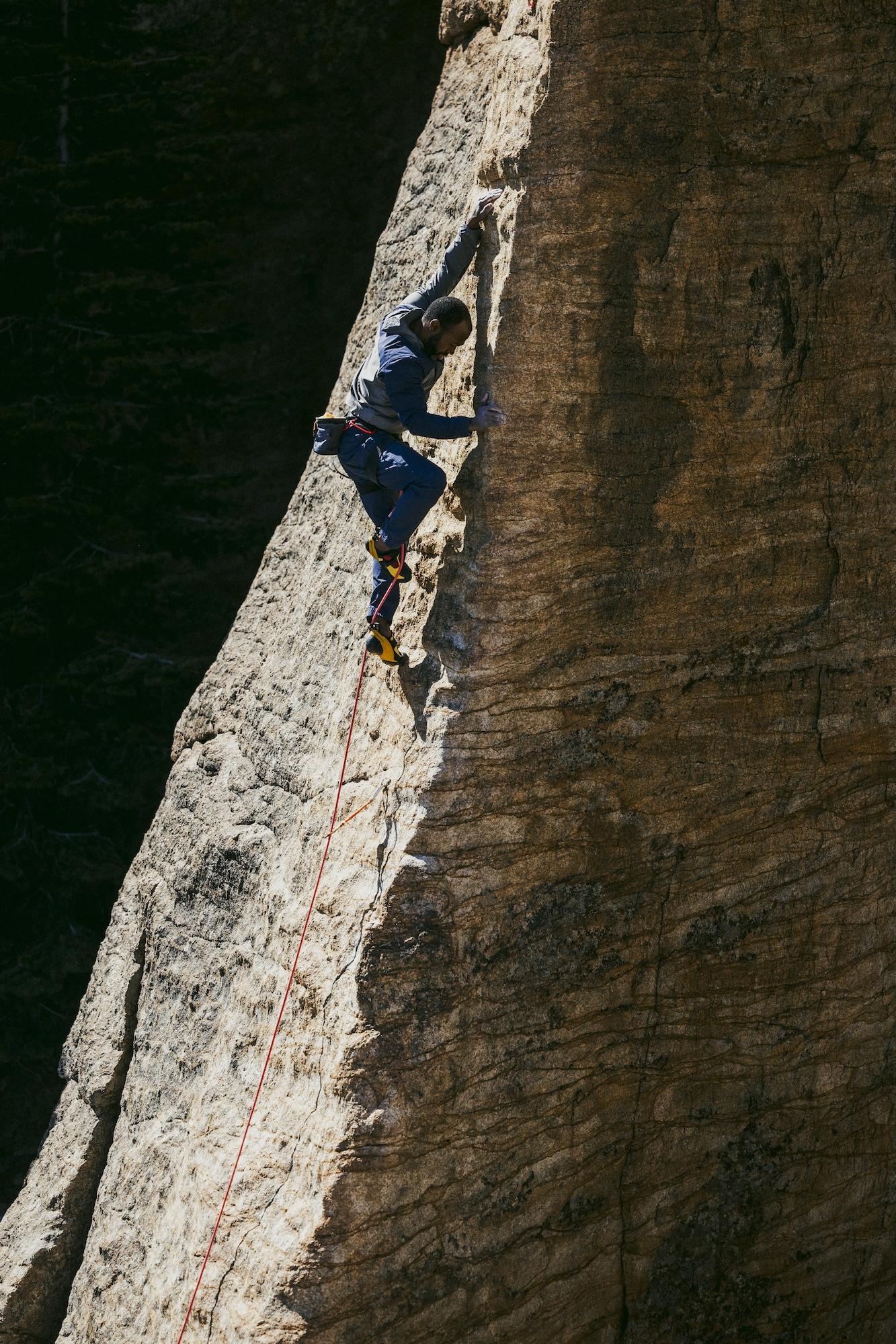 Climber scaling a steep, textured rock face with ropes in bright daylight.
