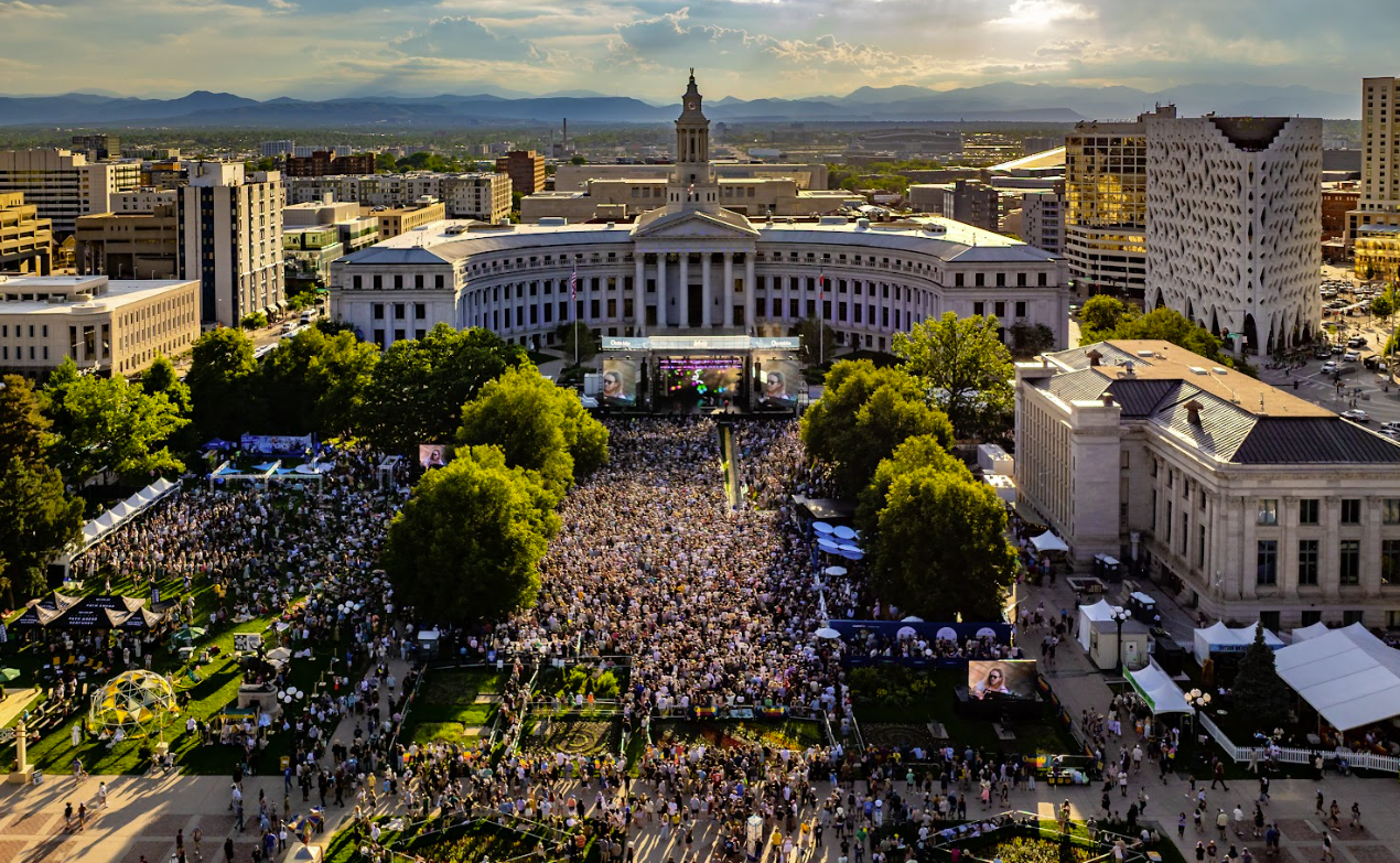 Crowd gathered in a park with a grand building and city skyline in the background.