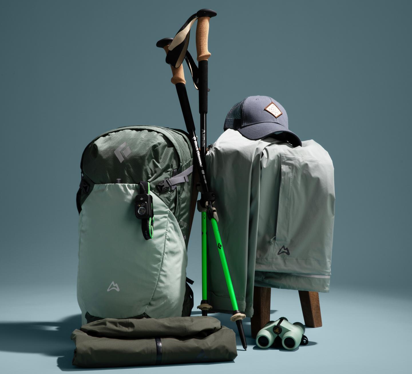Hiking gear setup featuring a backpack, jacket, cap, poles, and binoculars.