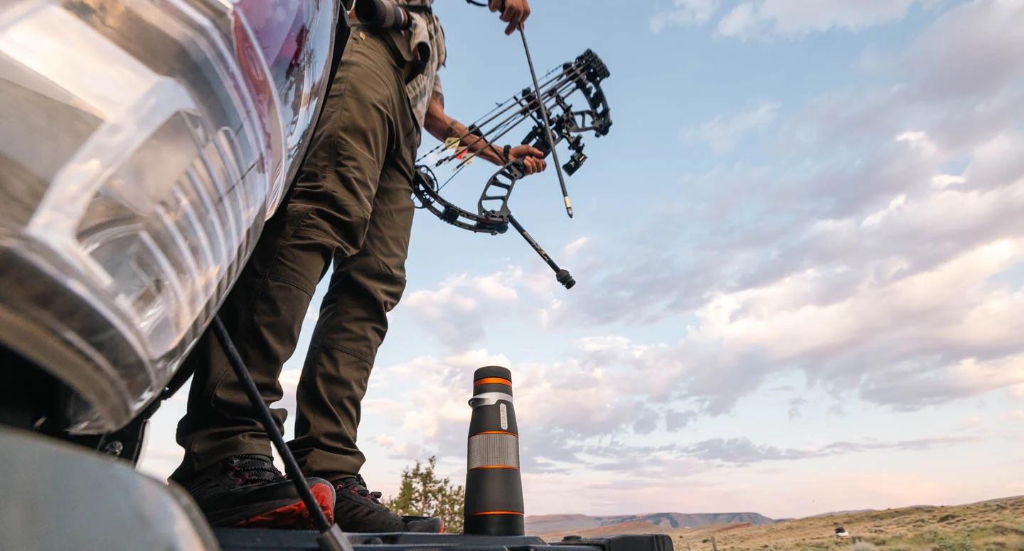 Person holds compound bow standing on truck; open landscape in background.