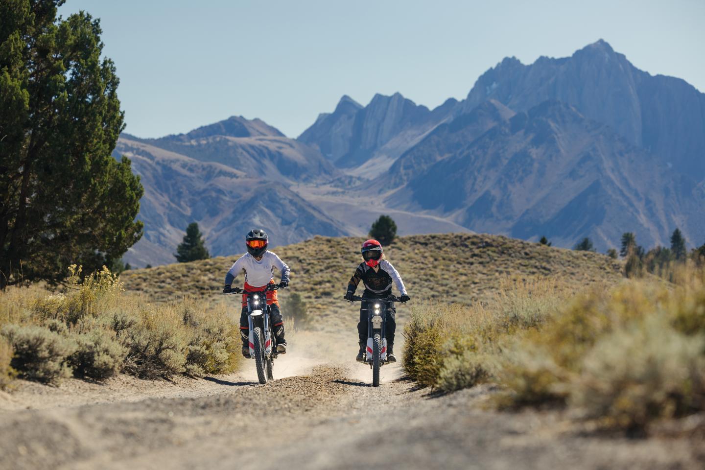 Two people riding electric dirt bikes on a doubletrack road with mountains in the background.