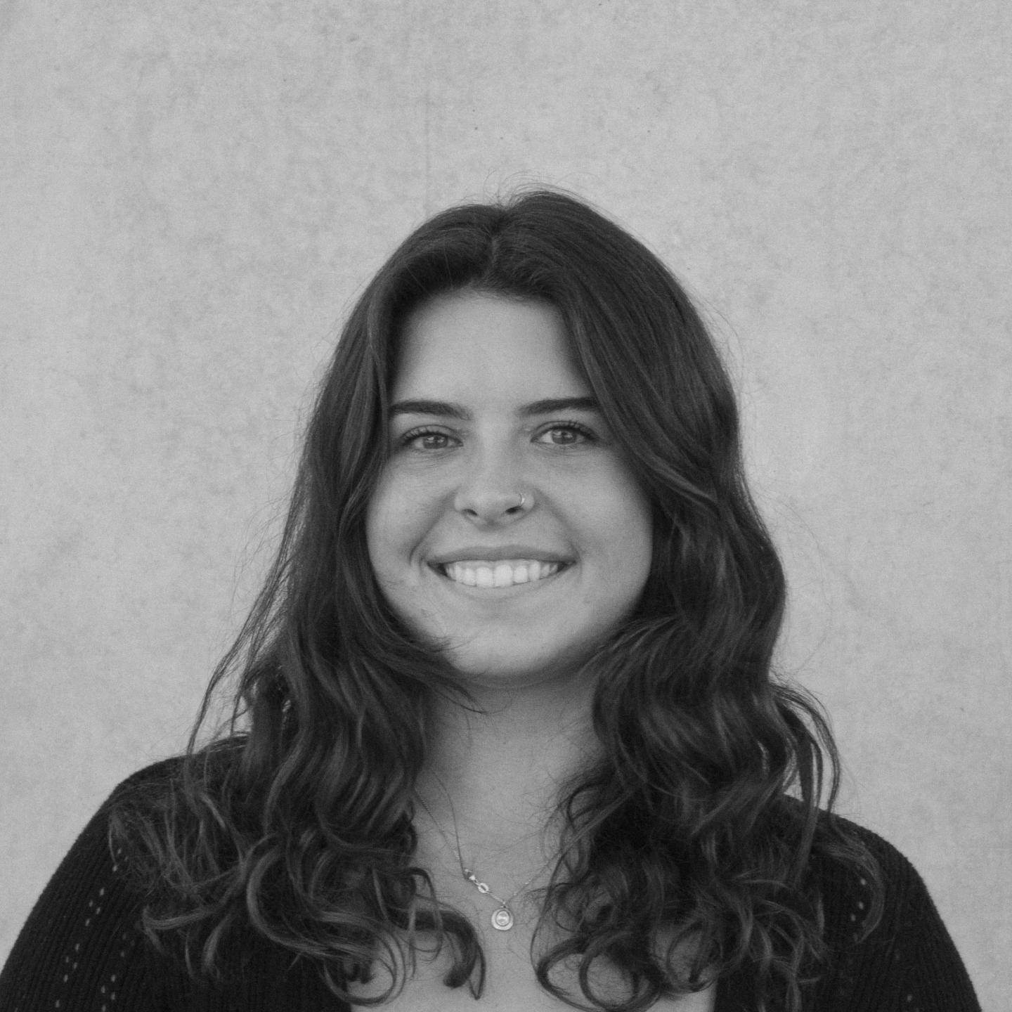 Smiling person with long wavy hair, wearing a necklace, against a plain background.