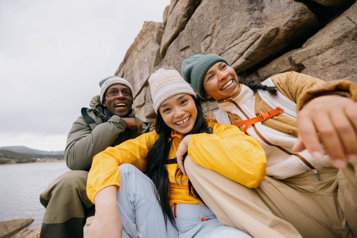 Three friends smile while sitting on rocks by a lake, wearing warm outdoor clothing.