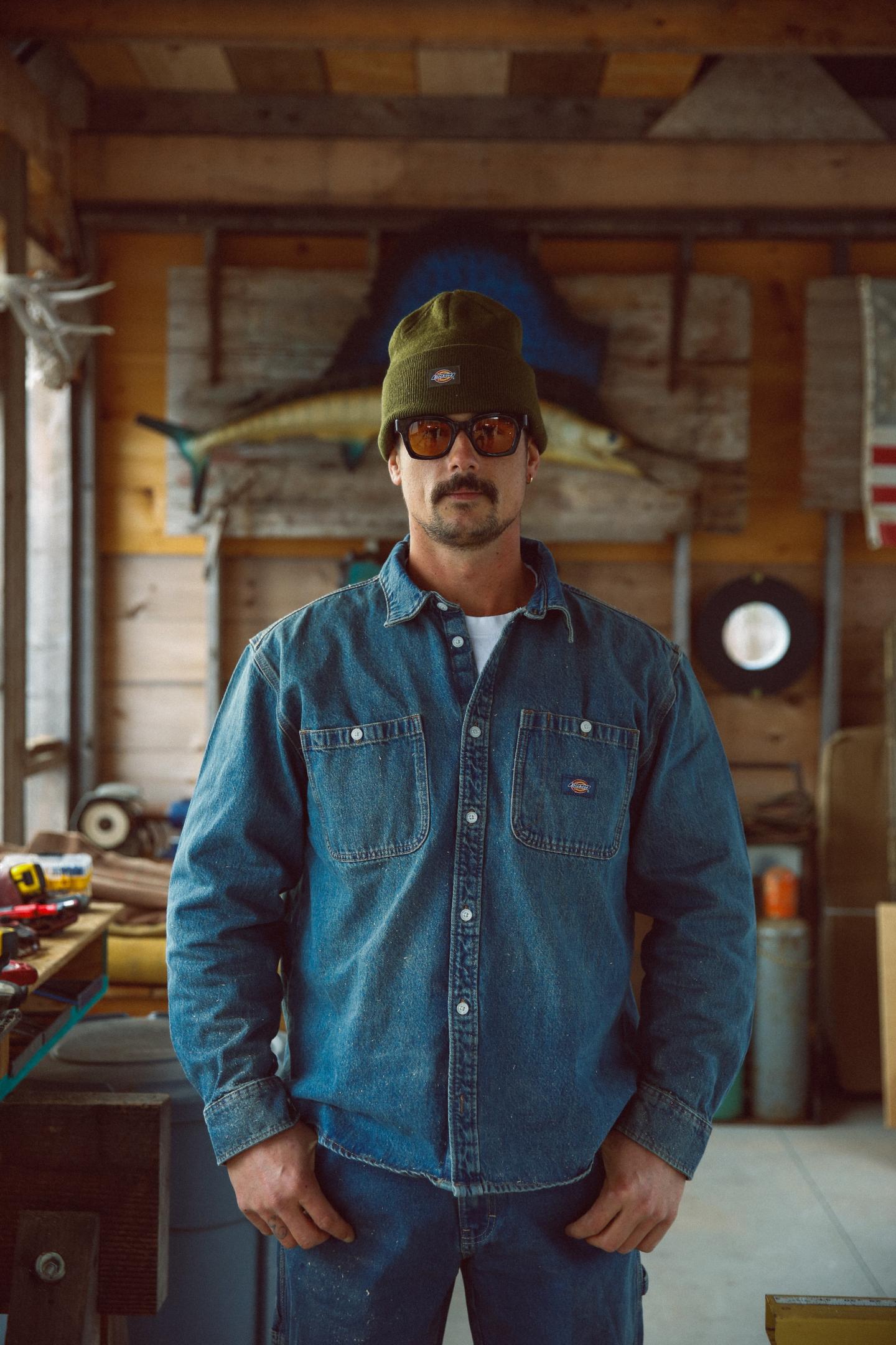 Man in denim shirt and beanie stands in rustic workshop.