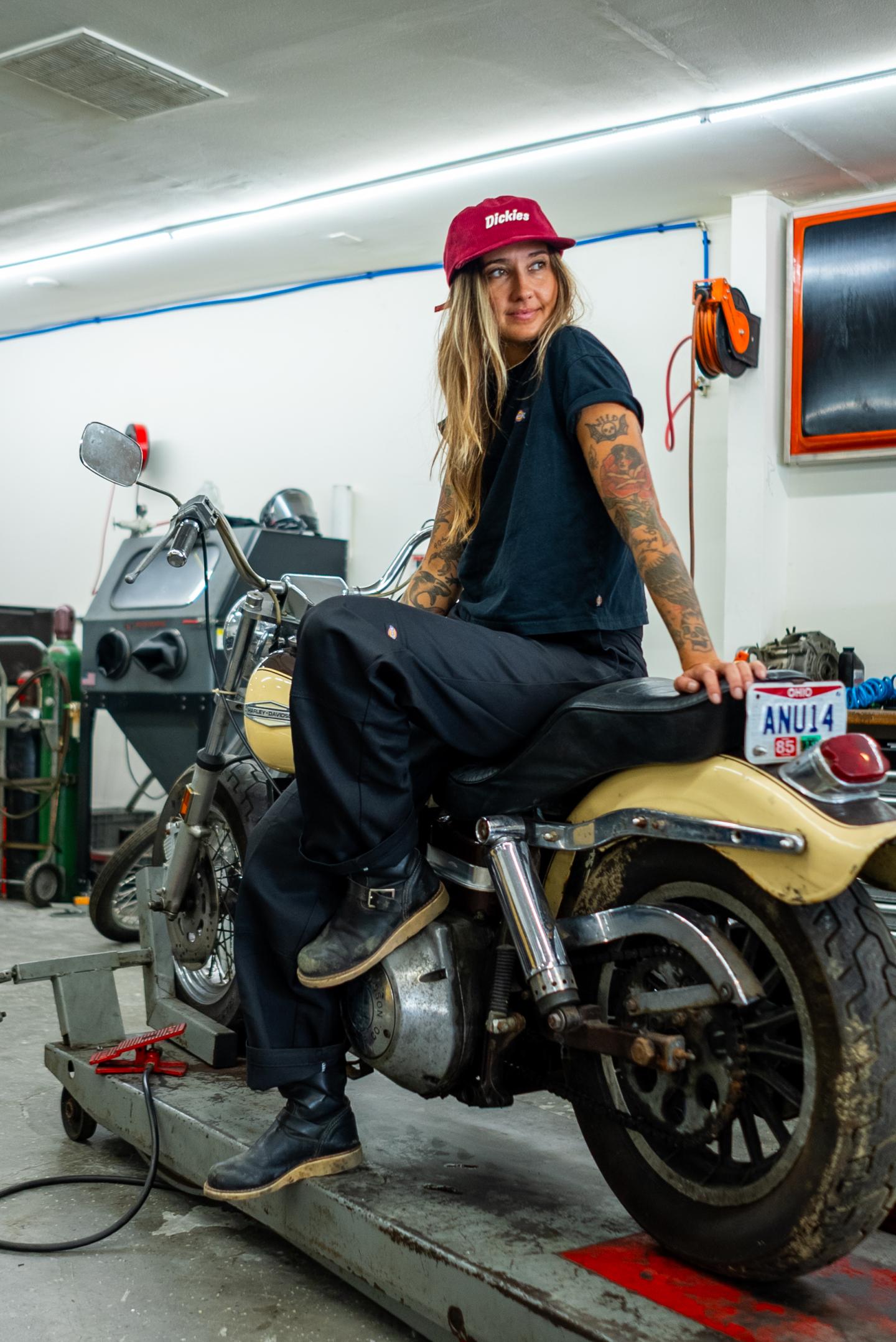 Person with tattoos sits smiling on a motorcycle in a garage, wearing a red cap and black outfit.