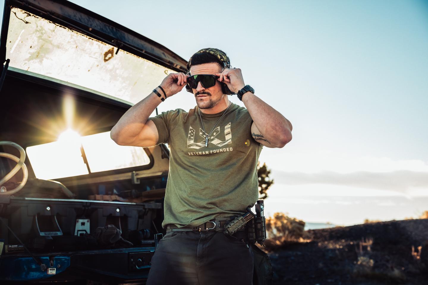Man adjusting WX Alpine sunglasses beside parked car at sunset.