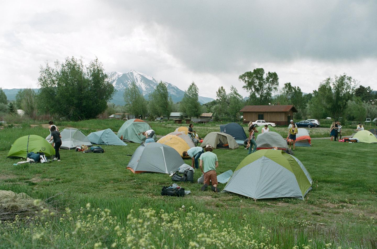 Camping scene with tents on a grassy field, mountains in the background.