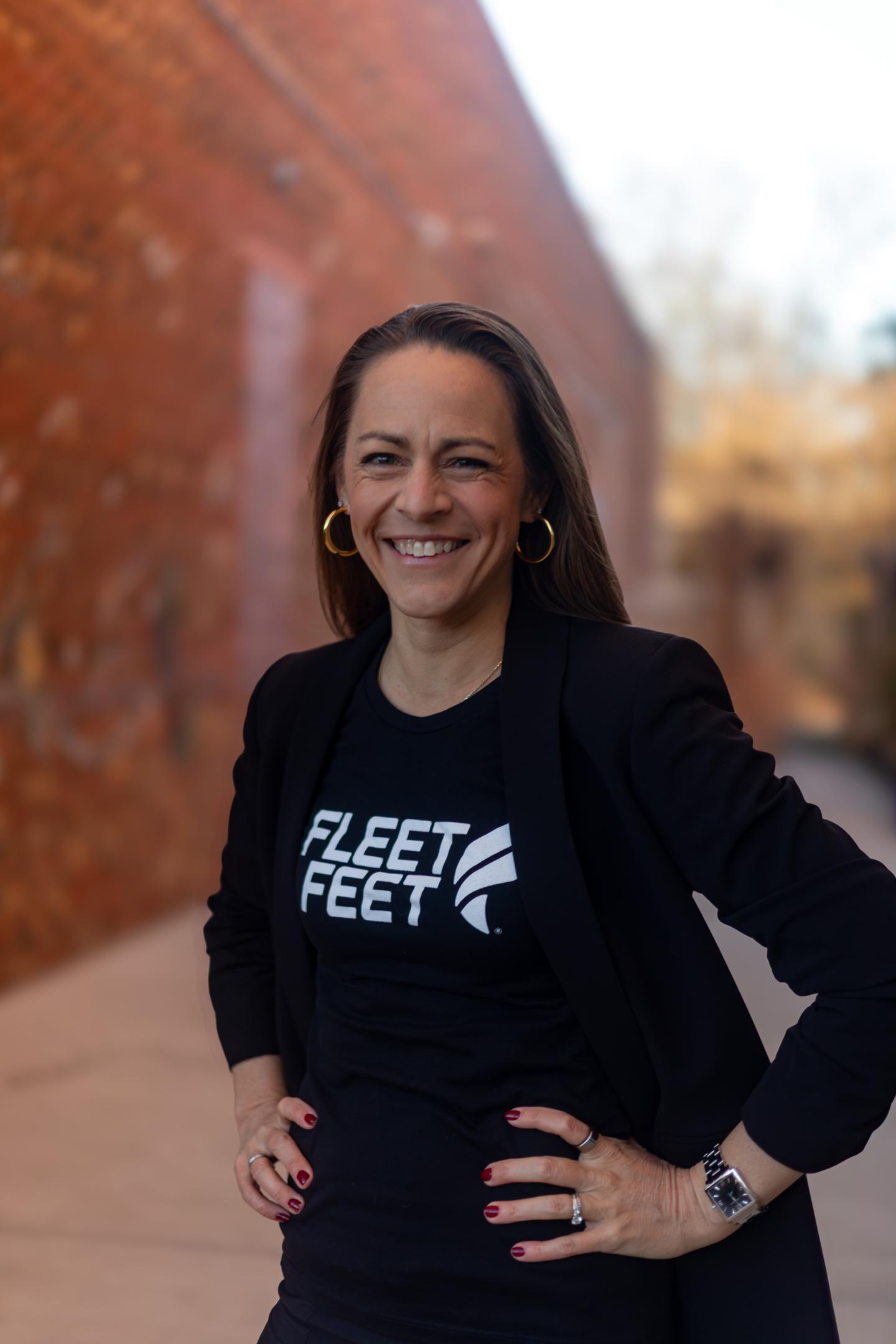 Smiling person in a black blazer outdoors against a blurred brick wall.