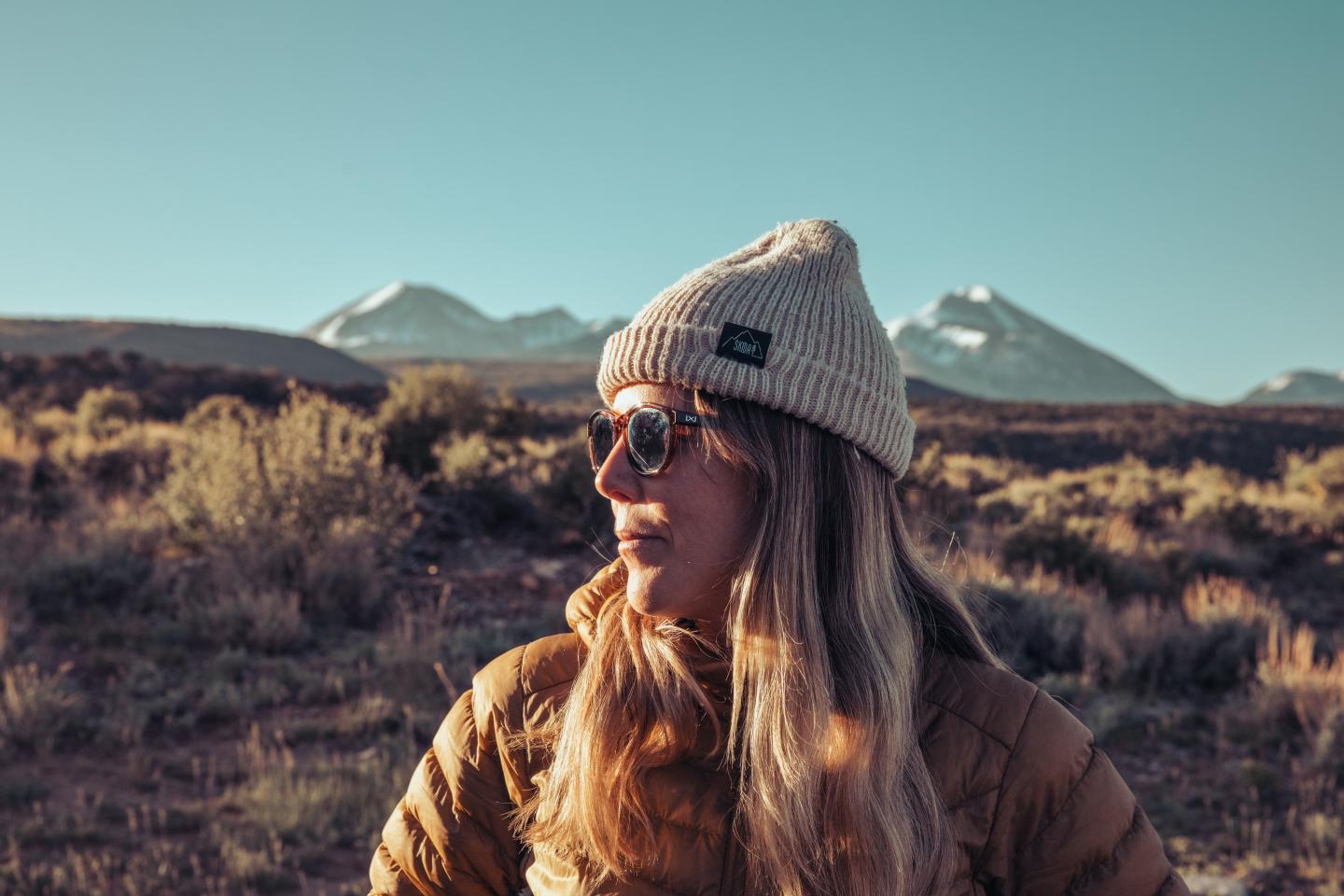 Person in a beanie and Wiley X sunglasses, with mountains in the background.