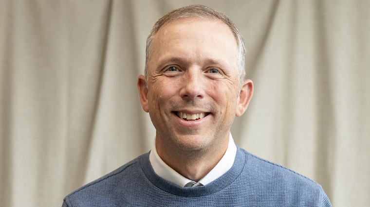 Smiling man in a blue sweater and collared shirt, neutral backdrop.