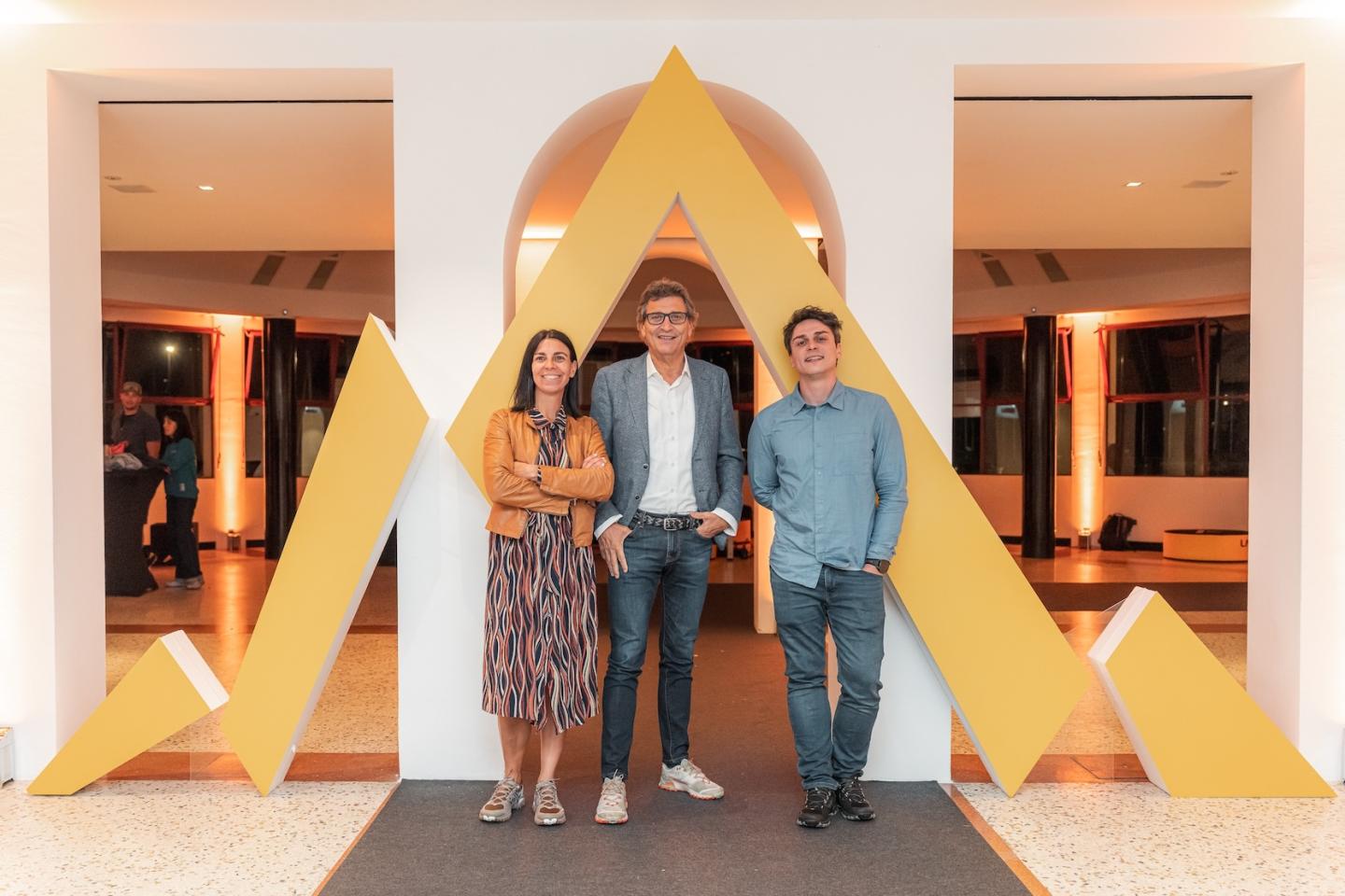 Three people standing in front of a large yellow triangular sculpture indoors.