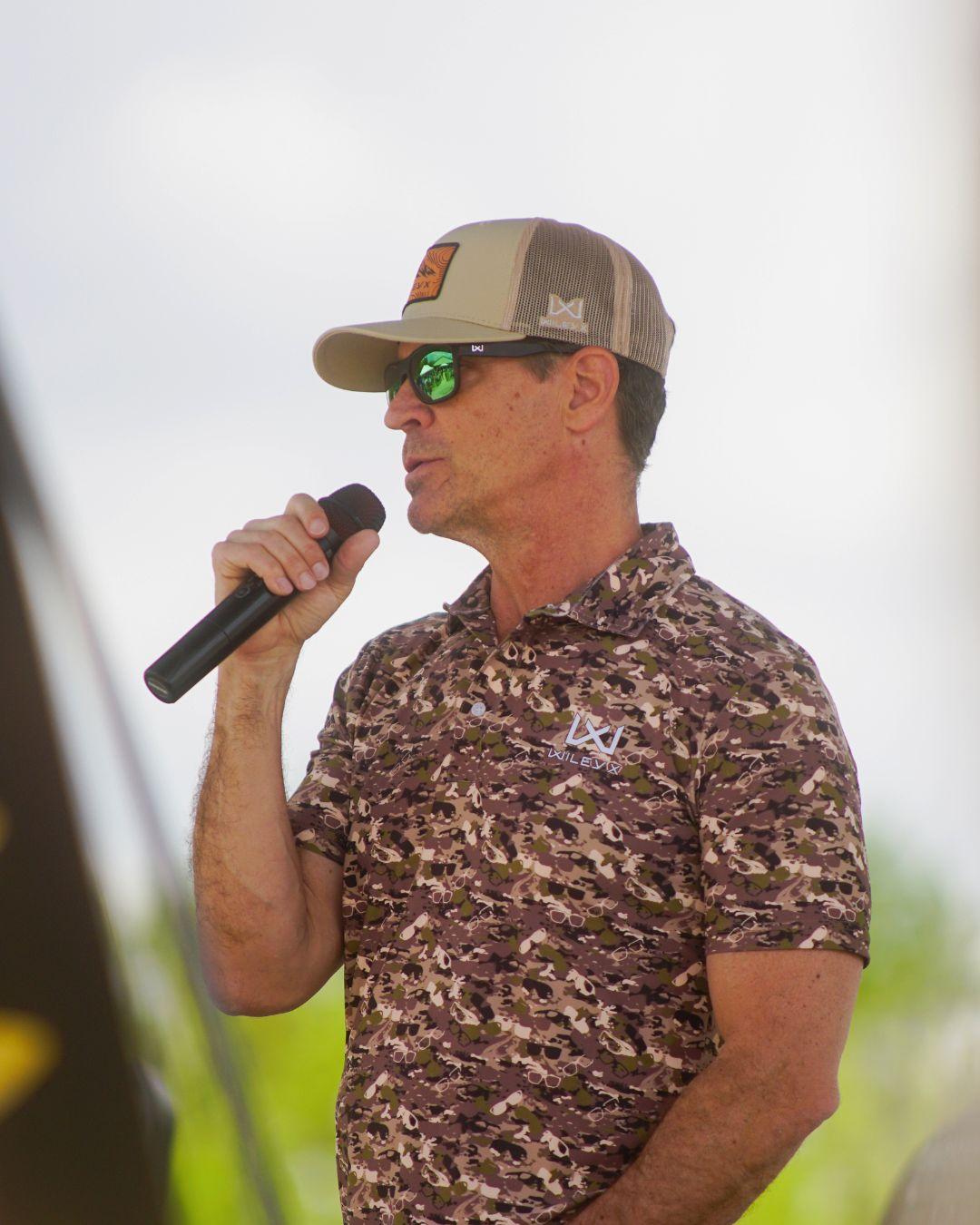 Man in camo shirt and cap speaks into a microphone outdoors.
