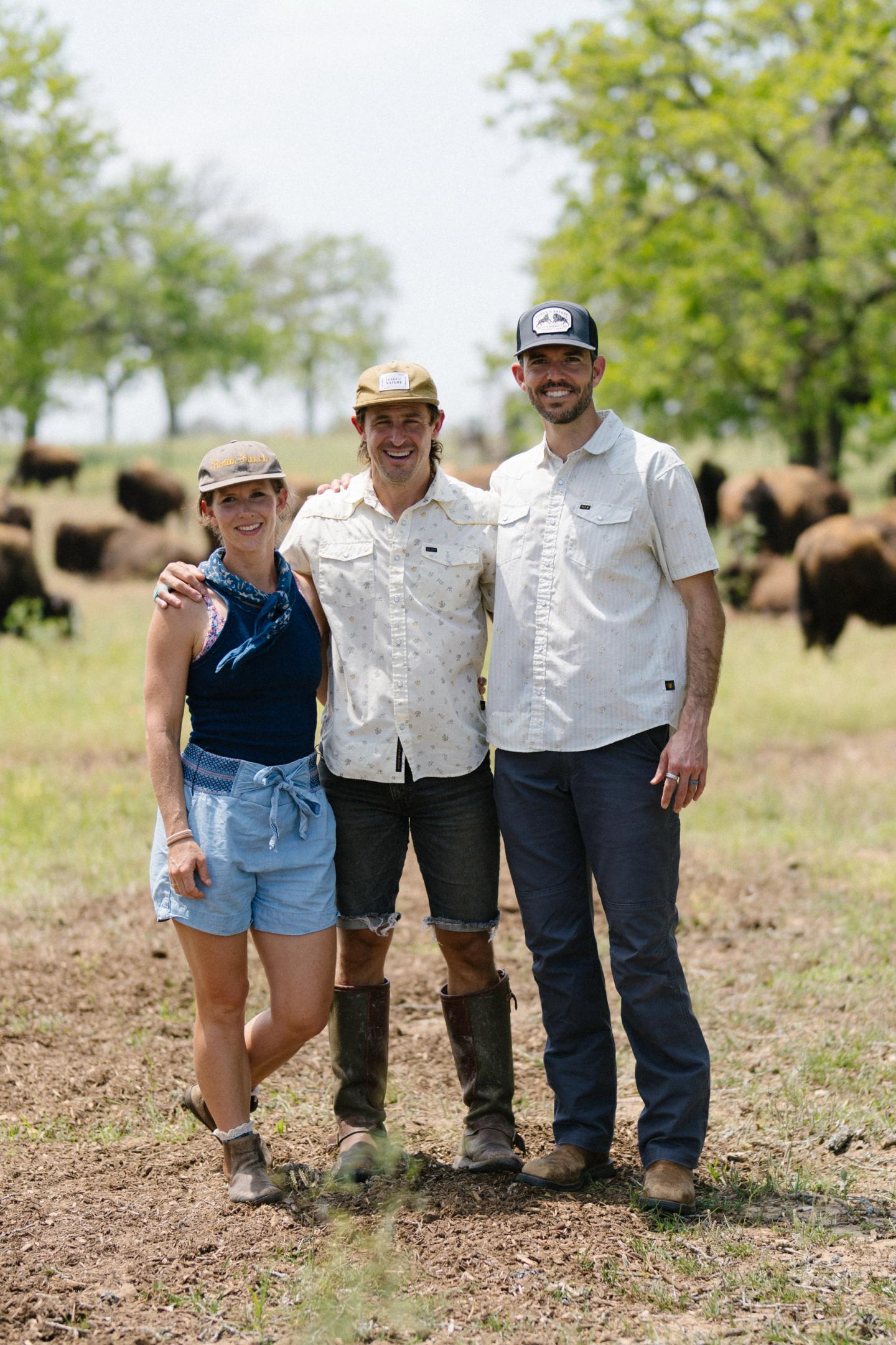 Three people smiling in a field with bison in the background.