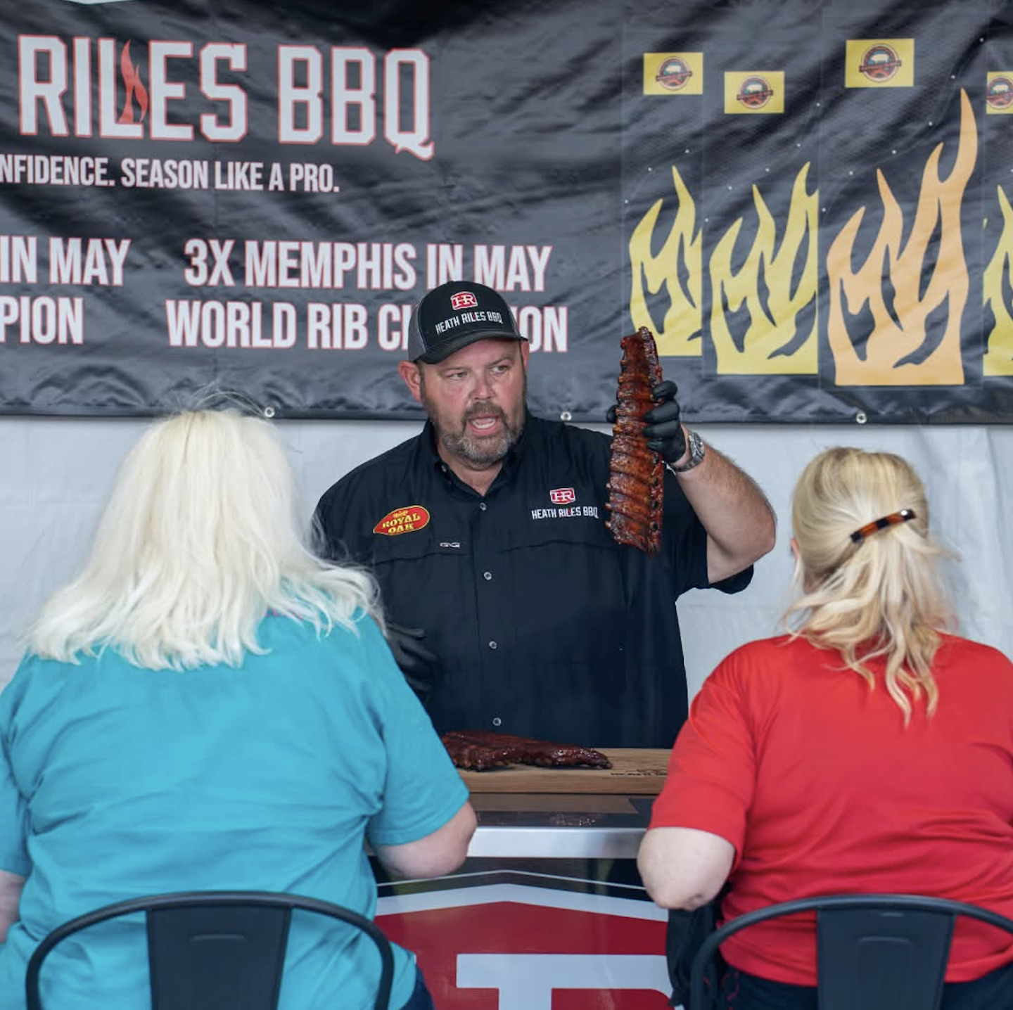Man holds ribs for two seated women at a BBQ event.