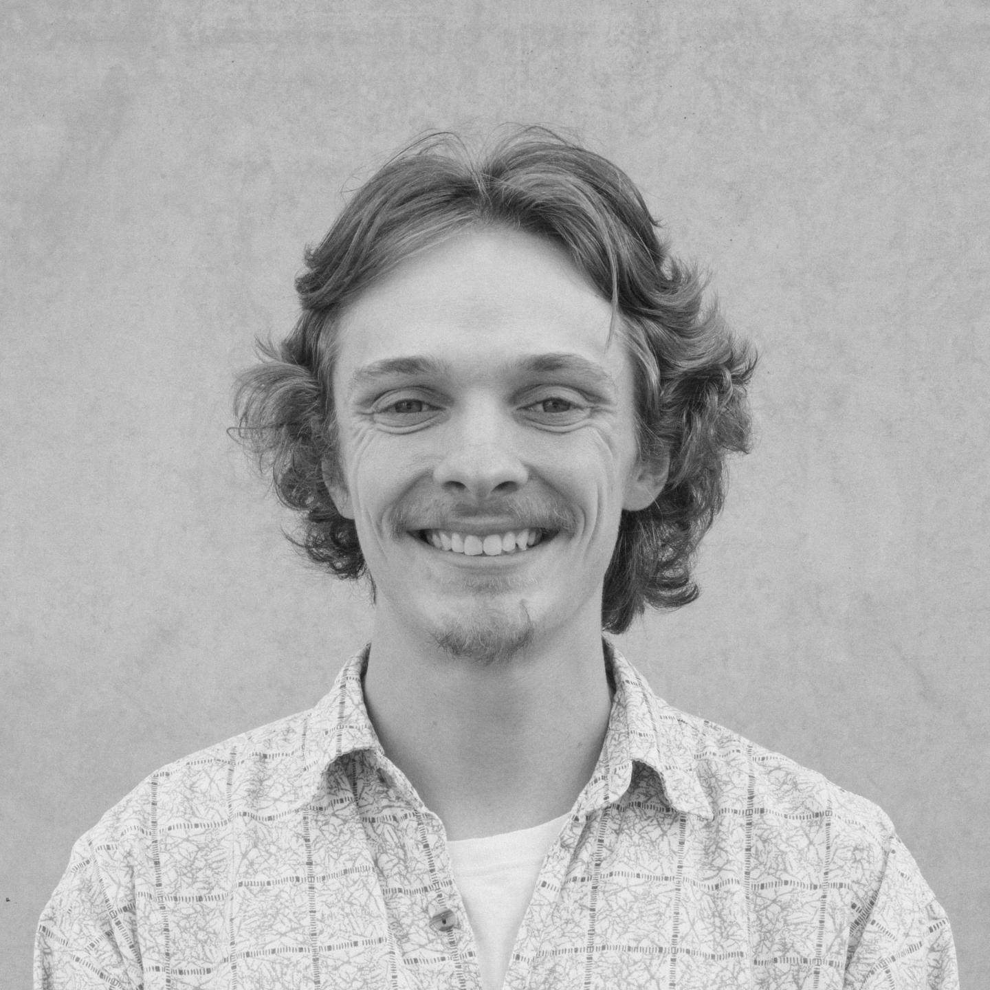Smiling person with curly hair, wearing a patterned shirt, in grayscale.
