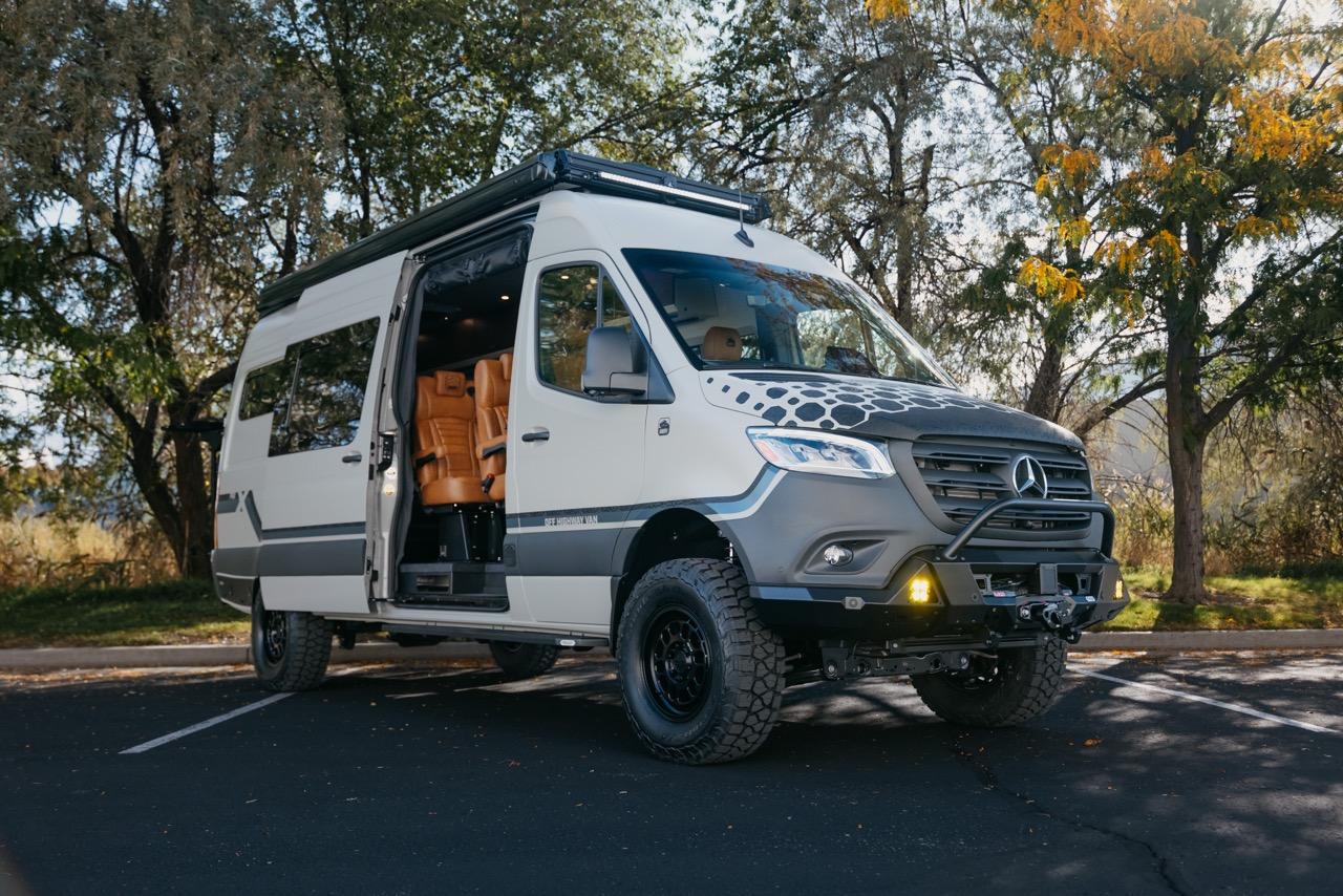 White camper van with open side door parked under autumn trees.